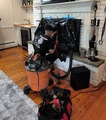 Person in protective clothing operating chimney-cleaning equipment indoors near a fireplace.
